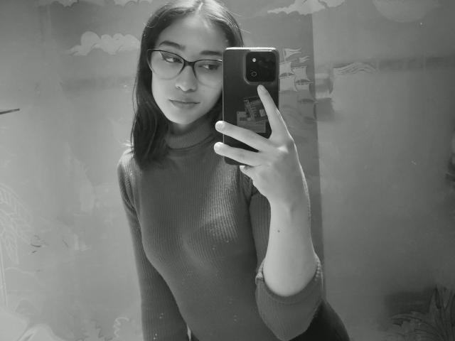 Autoportrait en noir et blanc de CamiVargas, portant des lunettes, avec une poitrine de taille moyenne, vêtue d'un pull col roulé texturé, tenant un téléphone, regardant légèrement sur le côté, avec un sourire discret..
