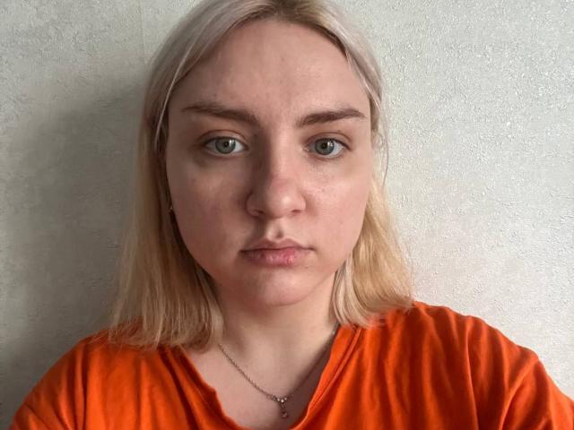 GlynisEchenique has pale skin, green eyes, and blonde hair. She's wearing an orange shirt with a silver necklace, looking serious with slightly parted lips.