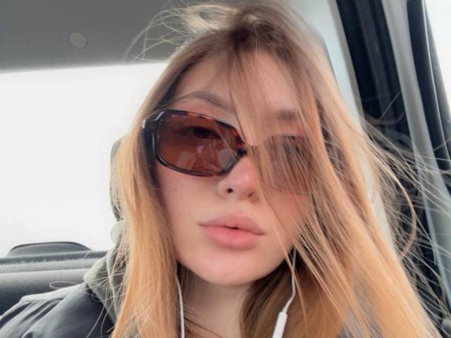 GigiWadkins has blonde hair, brown sunglasses, and white earbuds, with her pouty lips slightly parted, in a car, wearing a black hoodie.