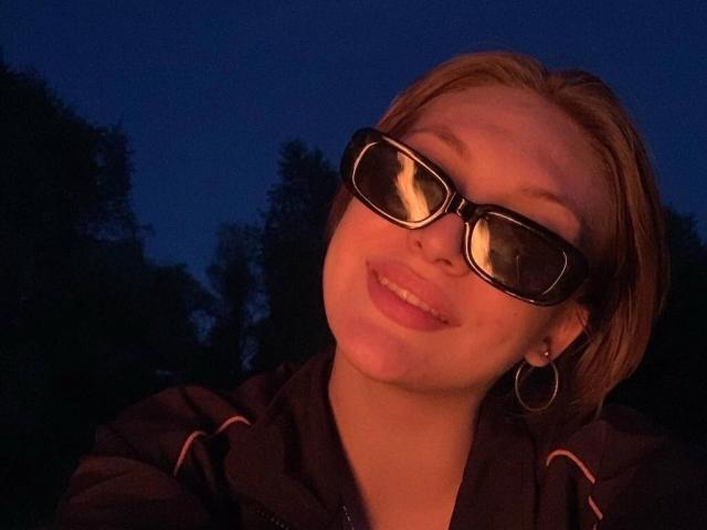 GigiWadkins with short brown hair, wearing black sunglasses and a dark hoodie, smiling at the camera, showing a small silver ear piercing on her left side.
