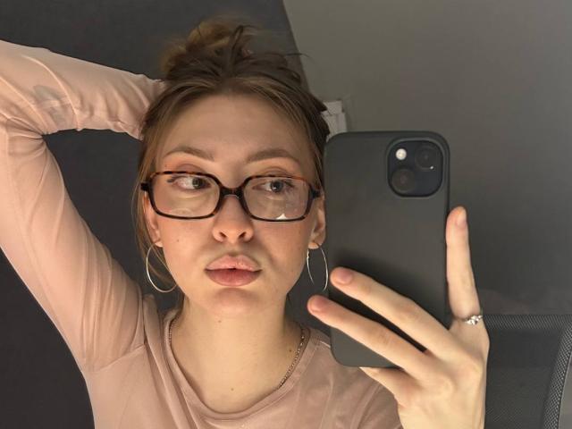 GigiWadkins with glasses, pink long-sleeve shirt, hoop earrings, ring, phone in hand, taking selfie, brown hair in bun, neutral expression, light skin, slight pout, casual look.
