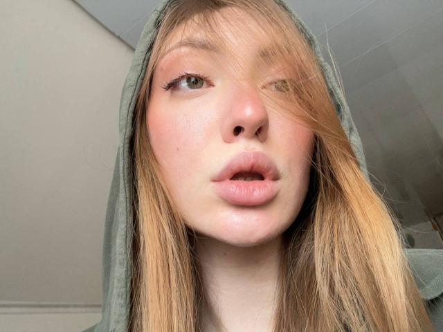 GigiWadkins has pouty pink lips, one eye winking, blonde hair in a green hood, fair skin, and subtle makeup with winged eyeliner. Her nose is small and cute.