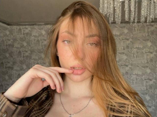 GigiWadkins has long, blonde hair, pinkish eyes, and a nose ring. She's biting her finger, wearing a brown plaid shirt, and a silver necklace.