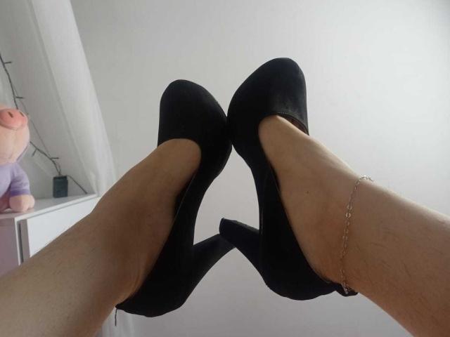 SophiaStorm's legs in black high heels, ankles adorned with a delicate gold chain, showing off her smooth, tan skin and slightly hairy calves, shot from a low angle.