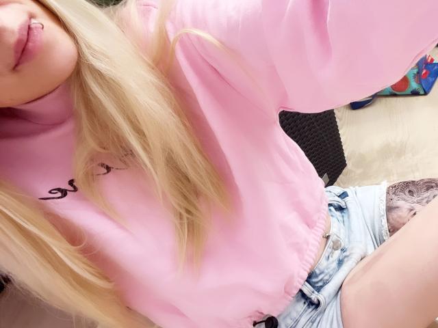 Lena, jeune et séduisante, avec des cheveux blonds, une chemise rose à manches longues et un short en jean, montrant son anneau rose sur la lèvre et le bord de sa culotte en dentelle. Ses seins, ses fesses et ses parties intimes sont partiellement visibles..