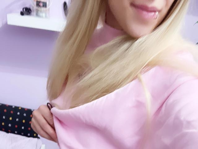 Lena, jung und attraktiv, mit blonden Haaren, einem pinken Oberteil, einem leichten Lächeln und einem Lippenpiercing, zeigt ihre kleinen Brüste, indem sie ihr Oberteil mit ihrer rechten Hand herunterzieht..