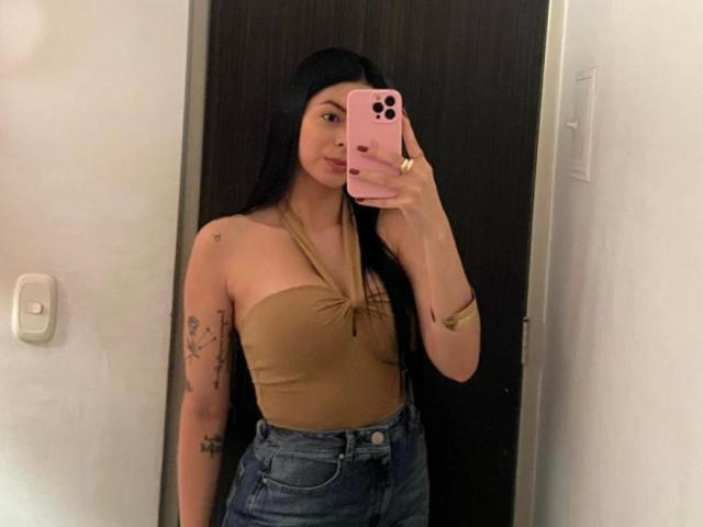 IsabellaJames with long black hair, tan strapless top, blue jeans, holding pink phone, showing tattoos on left arm, taking mirror selfie, showing her tits.