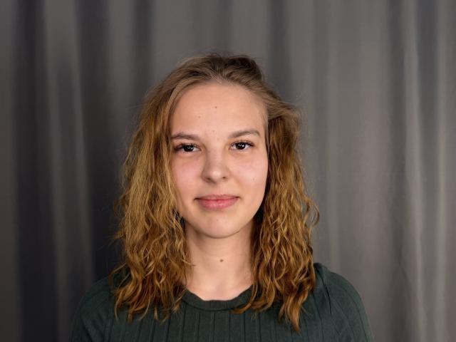 ChayaParrett heeft golvend, lichtbruin haar, een lichte huid en bruine ogen. Ze draagt een groene, geribbelde top en heeft een lichte glimlach op haar gezicht..