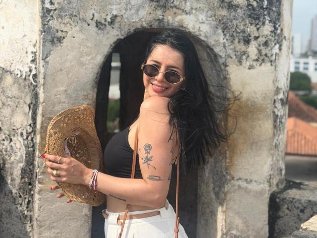 IsabellaJames with black tits in a black top, holding a straw hat, wearing round sunglasses, white pants, and red nails, has tattoos on her arm, smiling.