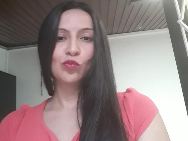 Erikka69 with long black hair, pink lips, and red shirt, pouting slightly, showing her nose and upper chest, standing in a room with a wooden ceiling.