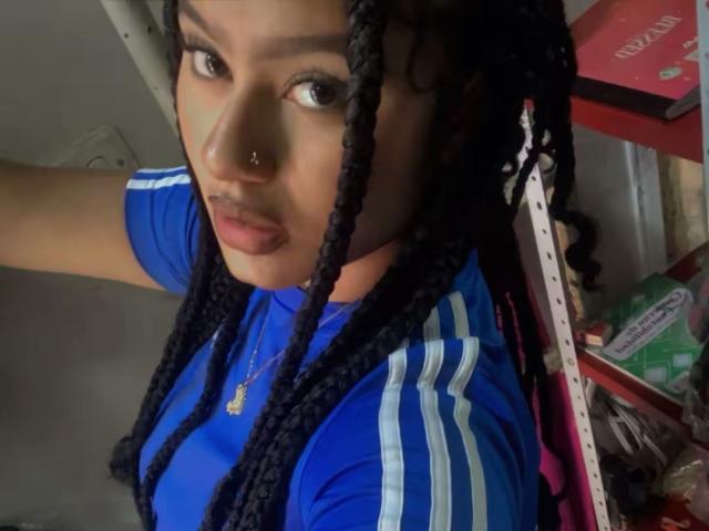 NoiraVale with black braided hair, blue Adidas shirt, nose ring, and gold necklace, looking up with pouty lips. Shelves with red and green boxes in the background.