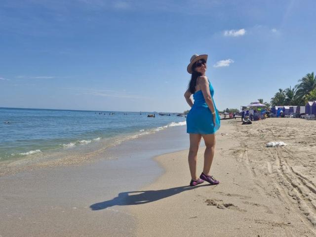 Erikka69 stands on a sandy beach in a blue dress, purple sandals, and a sunhat, with long dark hair and a tan butt, looking over the water.