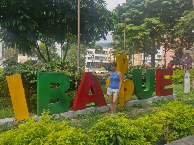 Erikka69 with blue top, white shorts, standing in front of colorful "I LOVE BAEU" letters, holding yellow "L". Green bushes, trees, and buildings in the background.