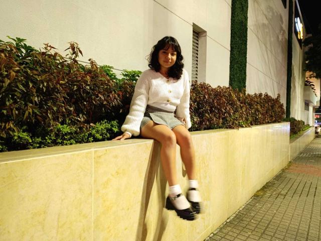 AsleyLoren, with curly black hair, sits on a yellow wall, wearing a white sweater, gray shorts, and black/white shoes, smiling at the camera, legs slightly apart.