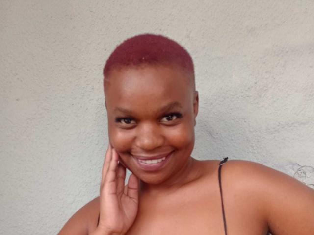 NastySquirtzX tiene la piel de color marrón oscuro, cabello corto teñido de rojo, y está sonriendo a la cámara con una mano cerca de su rostro, vistiendo una camiseta negra con tirantes finos..