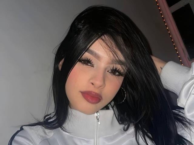 MilyLiams met zwart haar, lange wimpers, glanzende rode lippen, een witte jas met rits en oorringen, die naar de camera kijkt, met een hand in haar haar..