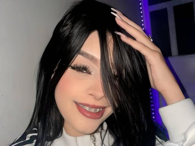 MilyLiams, avec des cheveux noirs, une chemise blanche à manches longues, et une lumière LED violette en arrière-plan, souriant, une main dans les cheveux, montrant des dents en métal et de longues ongles blancs..