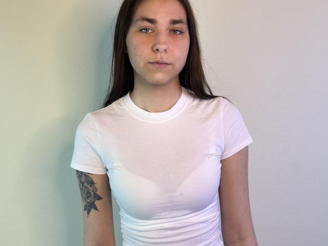 AlainaBlack with long brown hair, white shirt showing wet tits, small nipples, tattoo on right arm, standing against white wall, neutral expression, light skin, straight eyebrows.