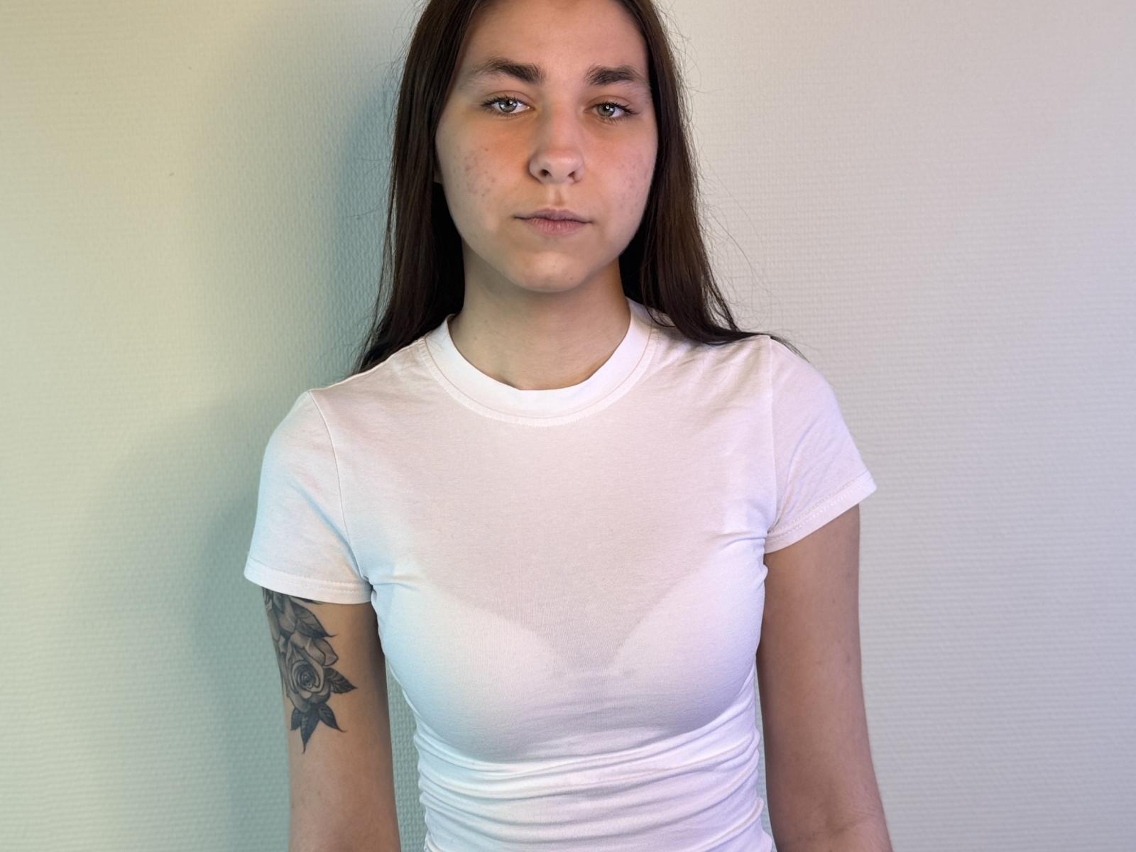 AlainaBlack, com cabelos castanhos longos, camisa branca revelando seios molhados, mamilos pequenos, tatuagem no braço direito, em pé contra uma parede branca, expressão neutra, pele clara, sobrancelhas retas..
