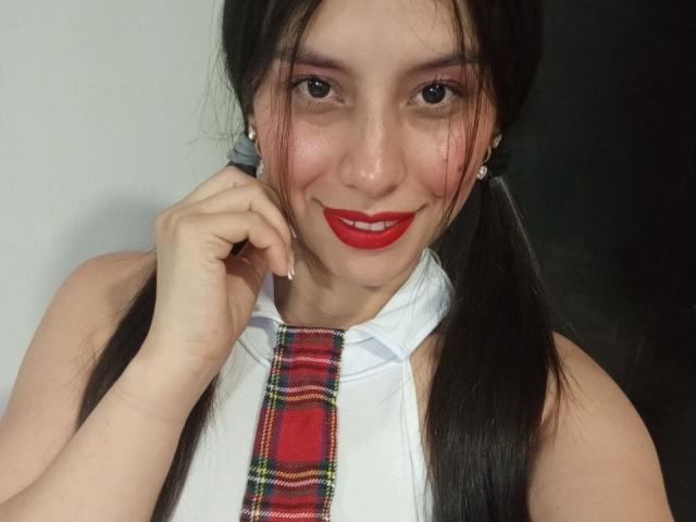 Madisson Fontaine, com maria-chiquinhas, vestindo uma camisa branca e gravata xadrez vermelha, tem batom vermelho, sombra vermelha e está sorrindo com a mão perto do rosto. Seus seios são pequenos..
