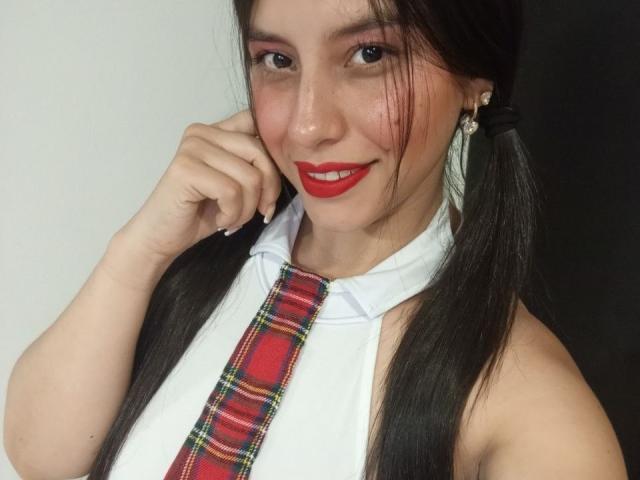 MadissonFontaine, com maria-chiquinhas, batom vermelho, blusa de alças branca e gravata xadrez vermelha, sorrindo, mostrando seus pequenos seios, segurando o queixo, cabelo escuro, pele clara e brincos..