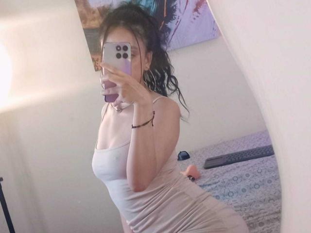 SharonKm in a tight, sheer, white dress, showing her tits, with wavy brown hair, taking a mirror selfie, holding a pink phone, in a softly lit room.