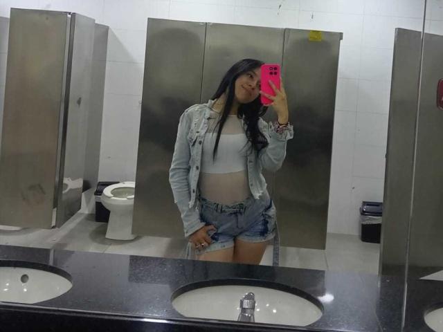 SharonKm with long black hair, wearing a white crop top, denim jacket, and blue shorts, taking a mirror selfie in a bathroom, holding a pink phone.