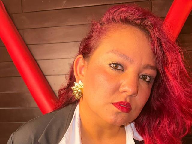 DorothyMoon with red hair, pouty lips, wearing red lipstick, gold earrings, and a grey suit, looking slightly annoyed, in a tight shot with red bars in the background.