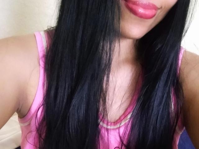 SoyLia, com cabelos longos e pretos, blusa de alça rosa e batom vermelho brilhante, mostrando parte do seu peito e rosto, com seus lábios brilhantes levemente entreabertos..