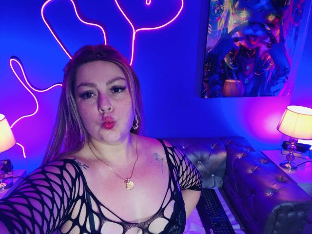 SamanthaGold, con labios rojos, expresión seria, vistiendo una blusa de malla negra que deja ver sus pechos, y un collar con forma de corazón, sentada en un sofá con capitonado, bajo luces de neón azules y rosas..