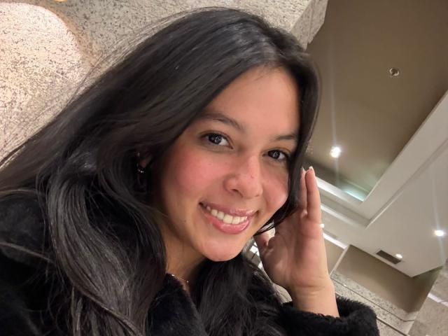 ValerieNoiir with dark, wavy hair, smiling, showing white teeth, wearing a black fur-like jacket, touching her ear, with brown eyes, and a slightly pink cheek.