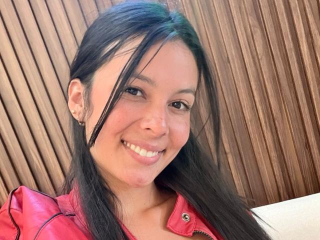 ValerieNoiir with long black hair, smiling, wearing a red jacket, has small earrings, and a slightly pinkish nose, in a selfie. Her tits are small, and she's cute.