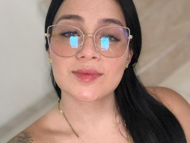 AnnyBentleii a la peau pâle, les cheveux noirs, et porte des lunettes à monture dorée qui reflètent un écran bleu. Elle sourit, ses lèvres sont roses, elle porte un collier en or et de petites boucles d'oreilles sont visibles..