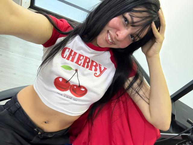 ValerieNoiir with long black hair, smiling, wearing a cherry rag tee, red sleeves, black pants, and a belly button piercing, hand in hair, seated on a chair.