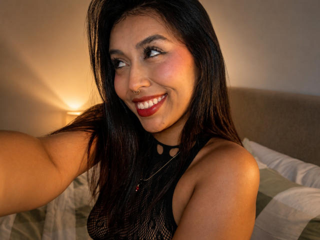 ZammySanz met donker haar, rode lippenstift, een neusring, lachend, draagt een zwarte kanten top, toont kleine borsten, maakt een selfie in de slaapkamer, heeft een lichtbruine huid, met warme verlichting..
