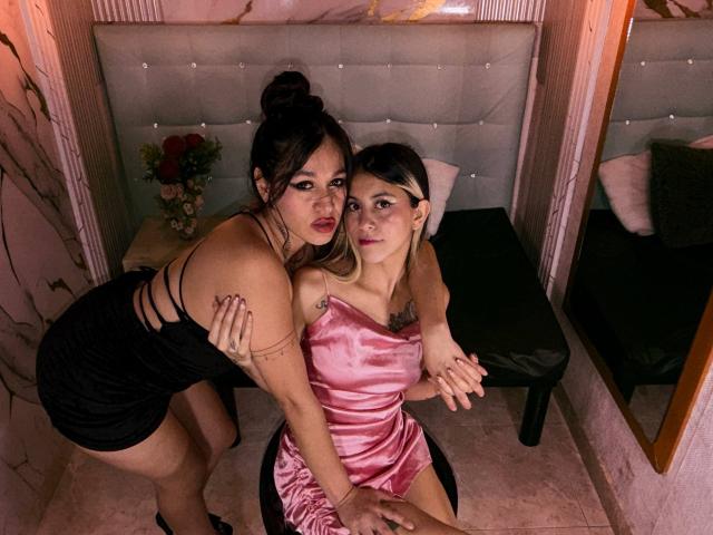AbbyNdYliria, con grandes senos, vestido negro y tatuajes, se inclina sobre AbbyNdYliria, que lleva un vestido de satén rosa y está sentada en una silla negra, tomadas de las manos, en una habitación con poca luz..