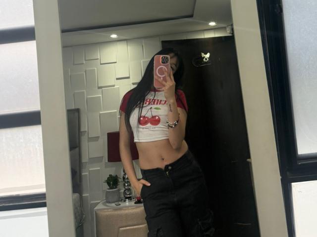 ValerieNoiir with long black hair, red and white crop top showing cherry print and small tits, black pants, navel piercing, holding pink phone, mirror selfie, hand in pocket.