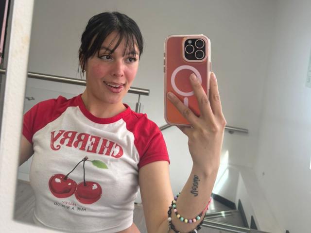 ValerieNoiir with dark hair, red and white ragged shirt, cherry print, holding phone, smiling, showing small tits, tattoo on right wrist, colorful bracelet, taking selfie in mirror.