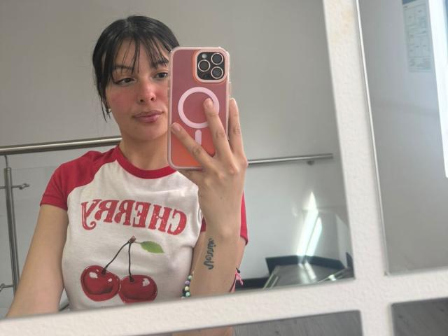 ValerieNoiir with dark hair in a white and red shirt with cherries and "YARD" text, holding a pink phone, showing a tattoo on her right wrist, mirror selfie.