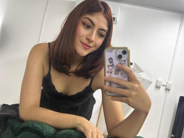 SpencerLe, avec des cheveux roux, un haut en dentelle noire, tenant un téléphone, souriant, montrant ses seins, un petit tatouage au poignet, une couverture verte, une porte blanche, un selfie pris devant un miroir, une coque de téléphone avec des autocollants..