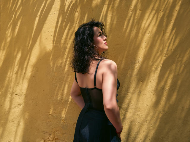 Elizabeth Dupont steht vor einer gelben Wand, trägt ein schwarzes, durchsichtiges Kleid, das ihren Hintern zeigt, hat welliges, dunkles Haar, und das Sonnenlicht wirft Schatten. Sie schaut nach oben, die Hände hinter dem Rücken..