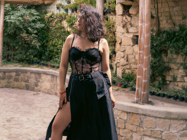 Elizabeth Dupont, mit lockigem, schwarzem Haar, trägt eine schwarze Spitzen-Bustière und einen schwarzen Rock mit einem seitlichen Schlitz, der ihren Oberschenkel und einen schwarzen Gürtel freigibt. Sie ist draußen und steht an einer Steinmauer..