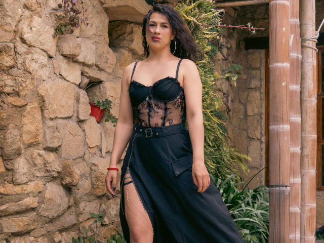 Elizabeth Dupont indossa biancheria intima di pizzo nero che mette in mostra il suo seno, e un'ampia gonna nera con una profonda spaccatura laterale, mentre è in piedi di fronte a un muro di pietra. Ha lunghi capelli scuri..