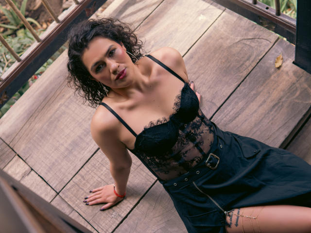 Elizabeth Dupont, avec ses cheveux noirs bouclés, portant un haut en dentelle noire et une jupe noire avec une ceinture, assise sur une terrasse en bois, regardant vers le haut avec un léger sourire..