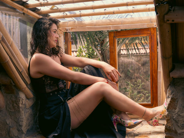 Elizabeth Dupont, con i capelli neri ricci, una camicetta di pizzo nero e una gonna nera, seduta in una finestra di legno, con le gambe incrociate, che mostra le cosce scoperte, tacchi con fiori e una posa rilassata, sexy e attraente..