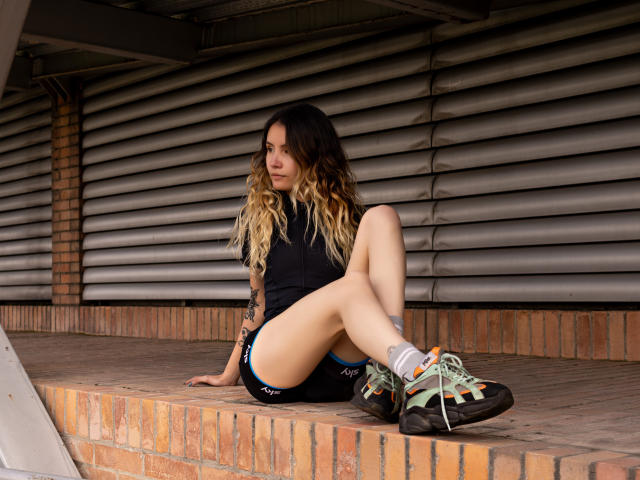 LilaLaurent, seduta con le gambe sollevate, ha un braccio tatuato, capelli biondi sfumati, una maglietta nera, pantaloncini neri e scarpe da ginnastica colorate, ed è appoggiata a un davanzale di mattoni, guardando di lato..