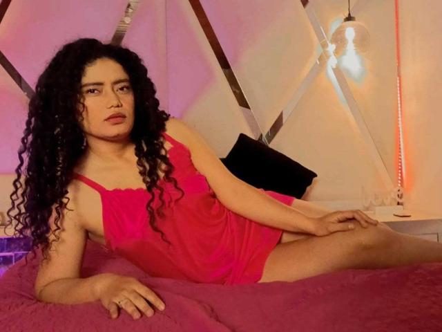 CurlyRuby with long curly black hair, wearing a pink dress, sitting on a pink bed, legs spread, right hand on bed, left hand on thigh, looking at camera.