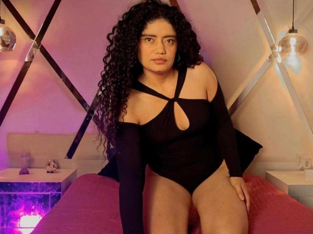 CurlyRuby, avec ses longs cheveux noirs bouclés, portant un haut noir à une épaule avec une découpe, assise sur un lit avec des draps roses, son derrière partiellement exposé, regardant la caméra..