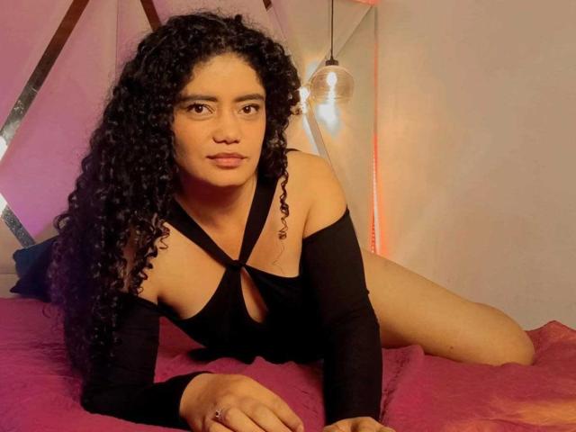 CurlyRuby, con i suoi capelli neri ricci, indossa una top nera con aperture, è sdraiata su un letto rosa, guarda la fotocamera, il suo sedere è appena coperto, e sullo sfondo c'è una lampadina..