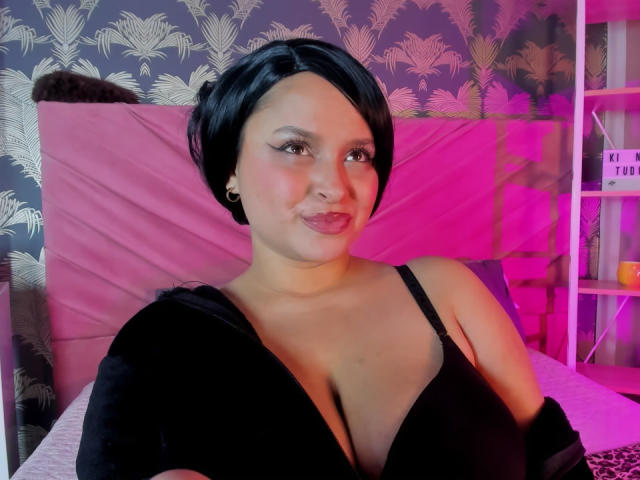 CoolPlay69 with short black hair, wearing a black dress, showing cleavage, smiling, pink and purple lighting, small hoop earrings, and a cute, confident look.